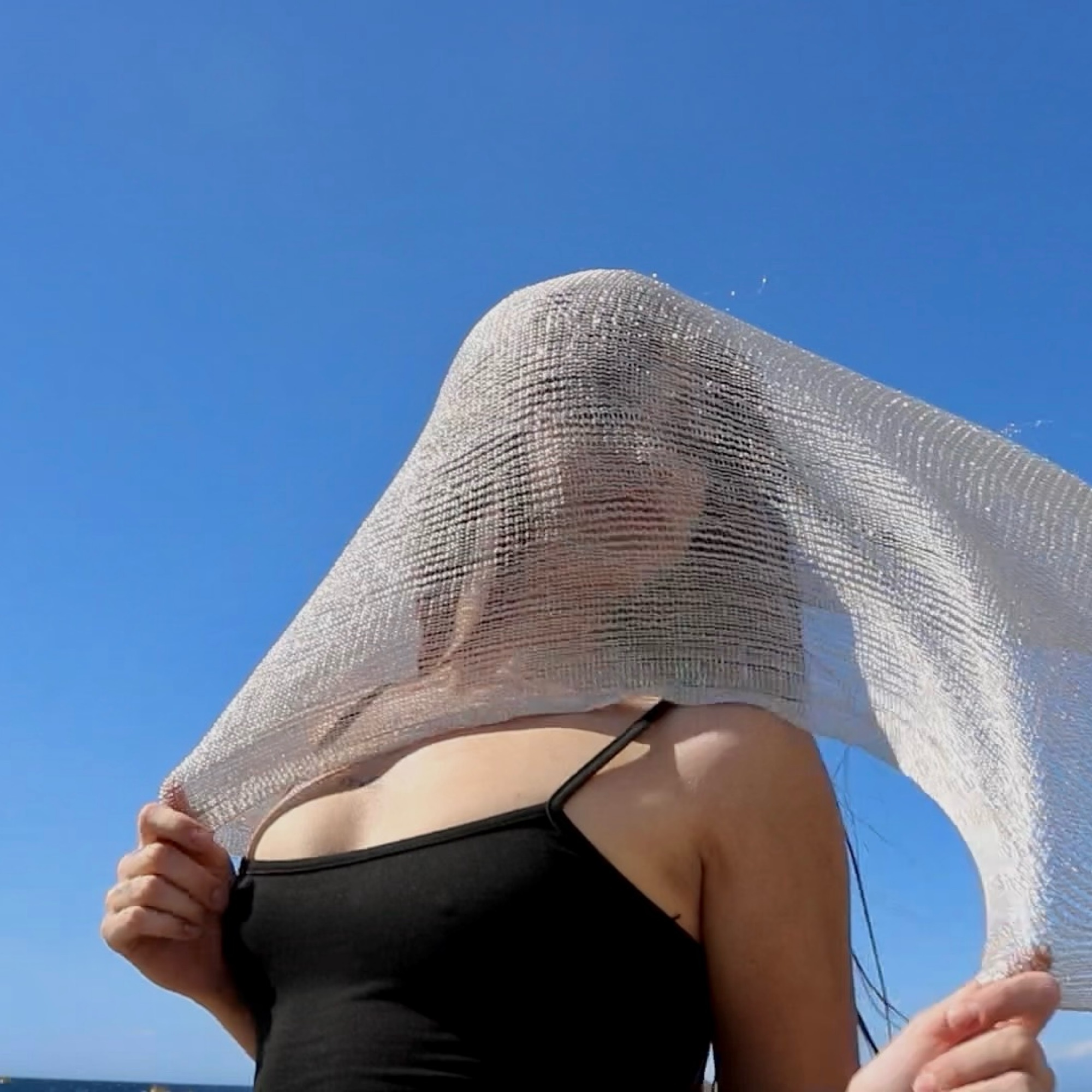 Catalina at the beach, a sheer sparkling fabric draped over her head and catching the sunlight, blue sky and sea behind her.